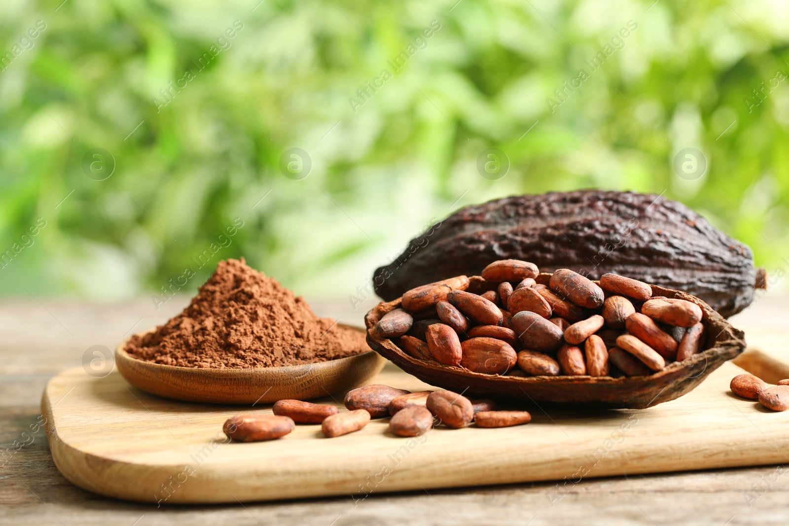 Photo of Board with cocoa pods, beans and powder on table against blurred green background