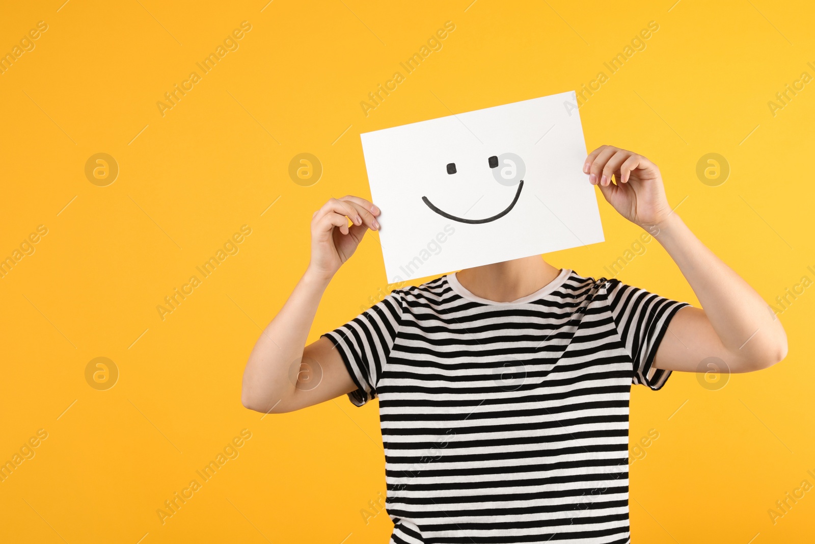 Photo of Woman hiding behind sheet of paper with happy face on yellow background, space for text
