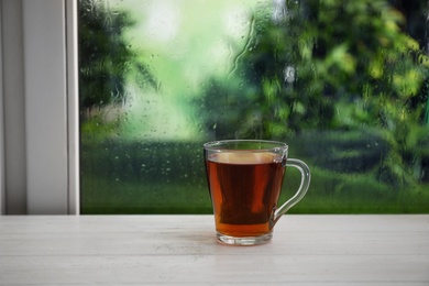 Photo of Cup of hot drink on wooden windowsill against glass with rain drops, space for text