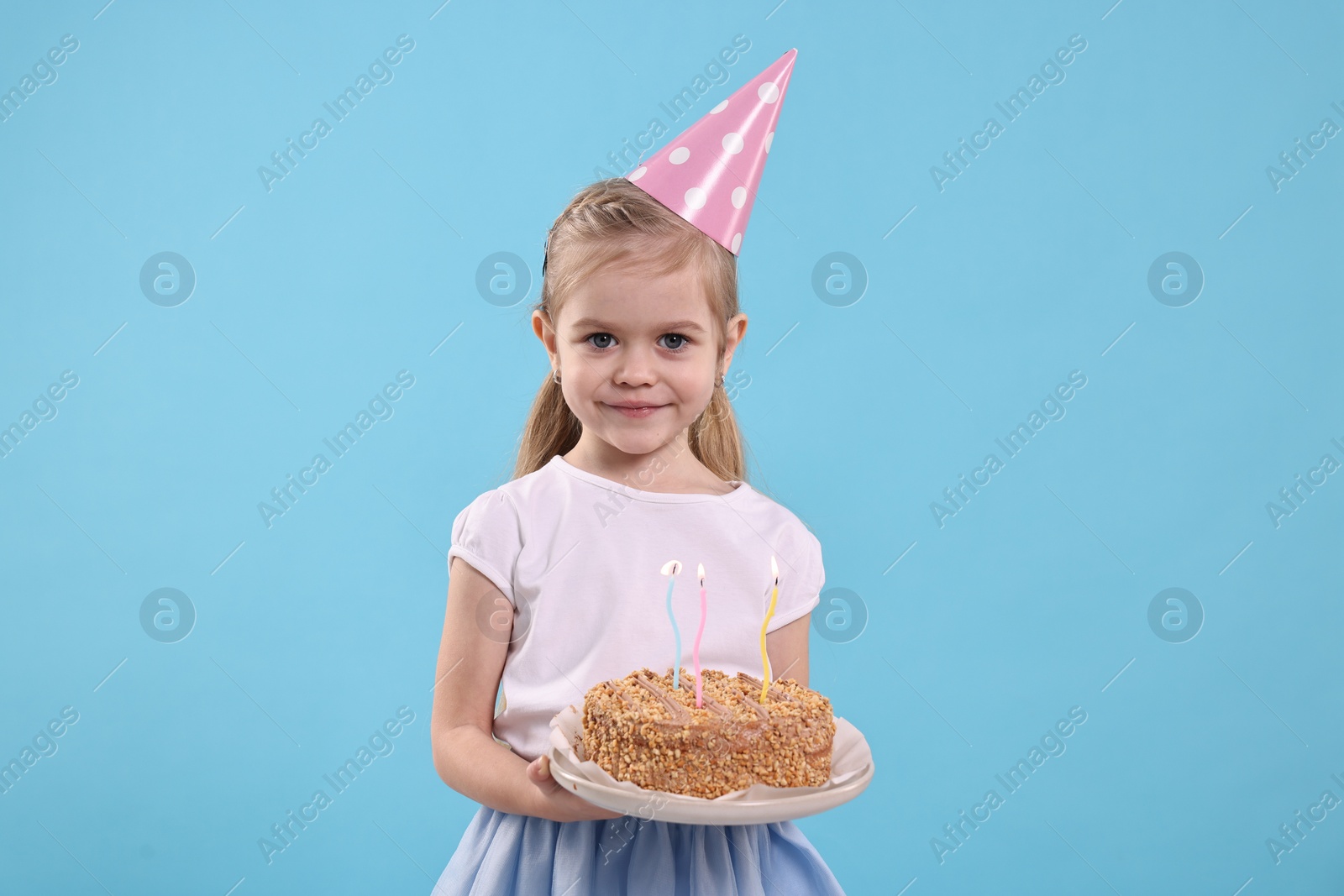 Photo of Birthday celebration. Cute little girl in party hat holding tasty cake with burning candles on light blue background
