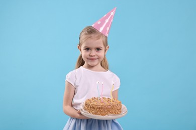 Birthday celebration. Cute little girl in party hat holding tasty cake with burning candles on light blue background