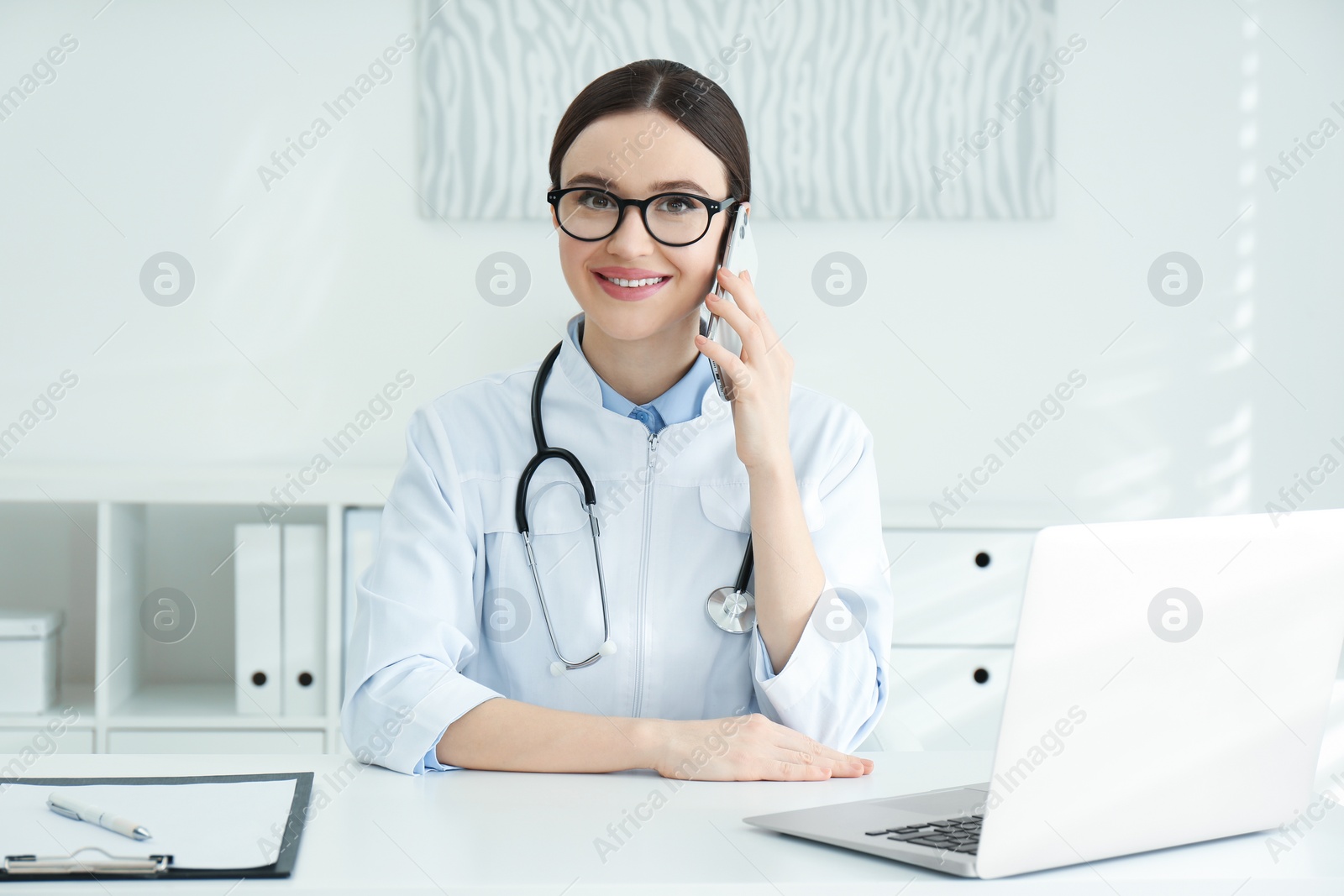 Photo of Young female doctor talking on phone at table in office