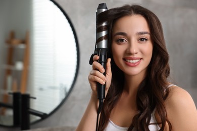 Photo of Smiling woman using curling hair iron in bathroom. Space for text