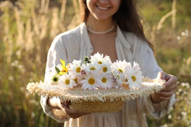 Photo of Woman holding straw hat with beautiful wild flowers outdoors, closeup
