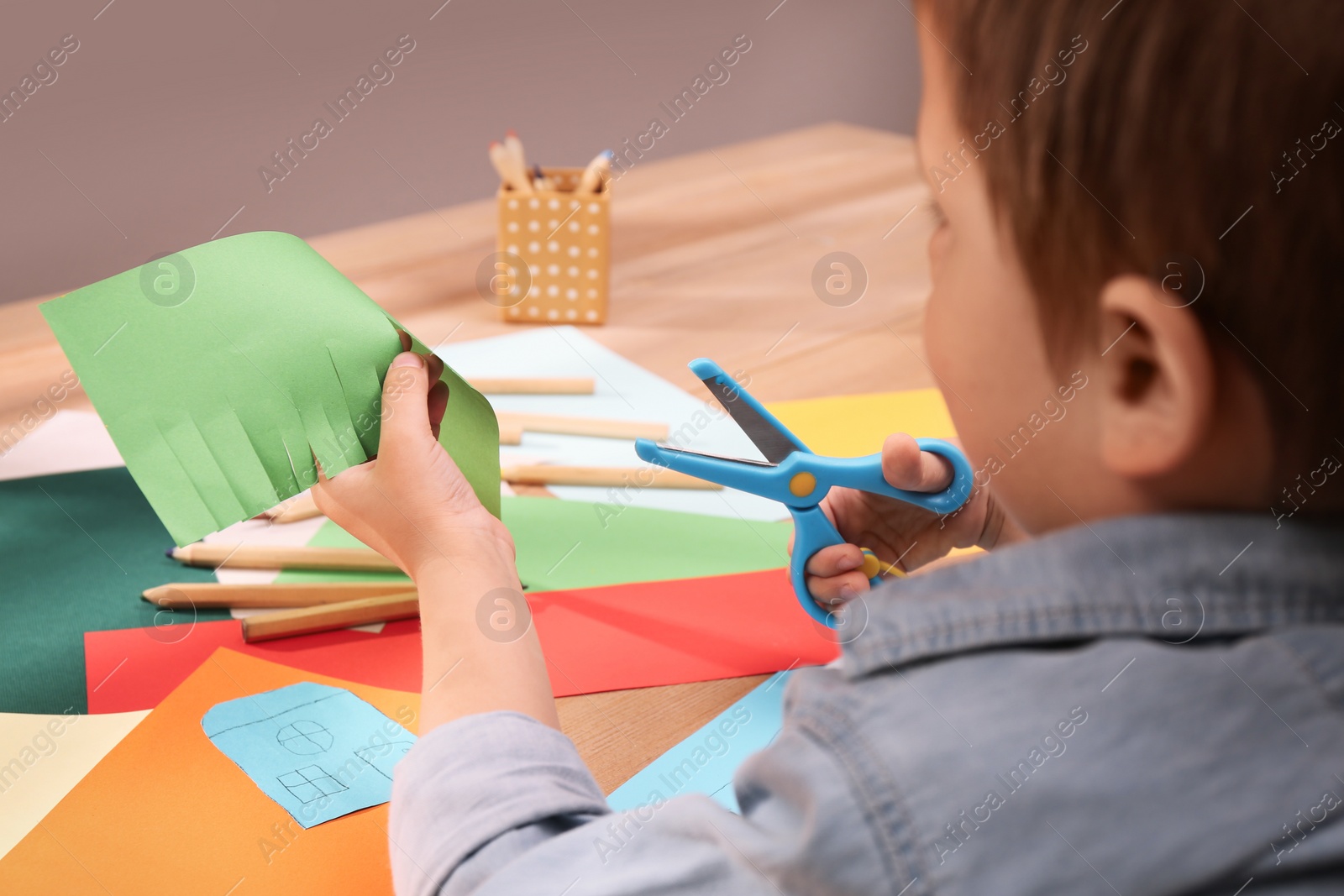 Photo of Little boy cutting color paper with scissors at table indoors, closeup