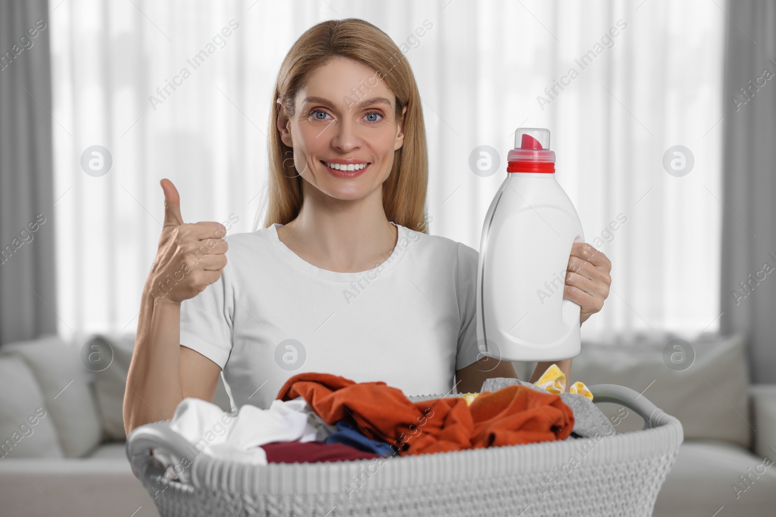 Photo of Woman holding fabric softener near basket with dirty clothes and showing thumbs up in room