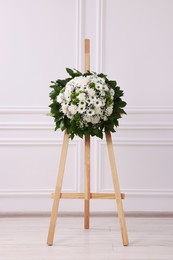 Photo of Funeral wreath of flowers on wooden stand near white wall indoors