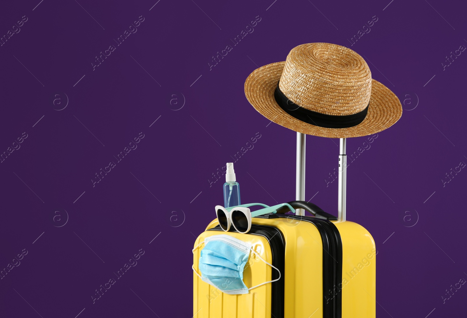 Photo of Stylish yellow suitcase with protective mask, sunglasses, hat and antiseptic spray on purple background, space for text. Travelling during coronavirus pandemic