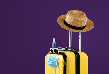 Stylish yellow suitcase with protective mask, sunglasses, hat and antiseptic spray on purple background, space for text. Travelling during coronavirus pandemic