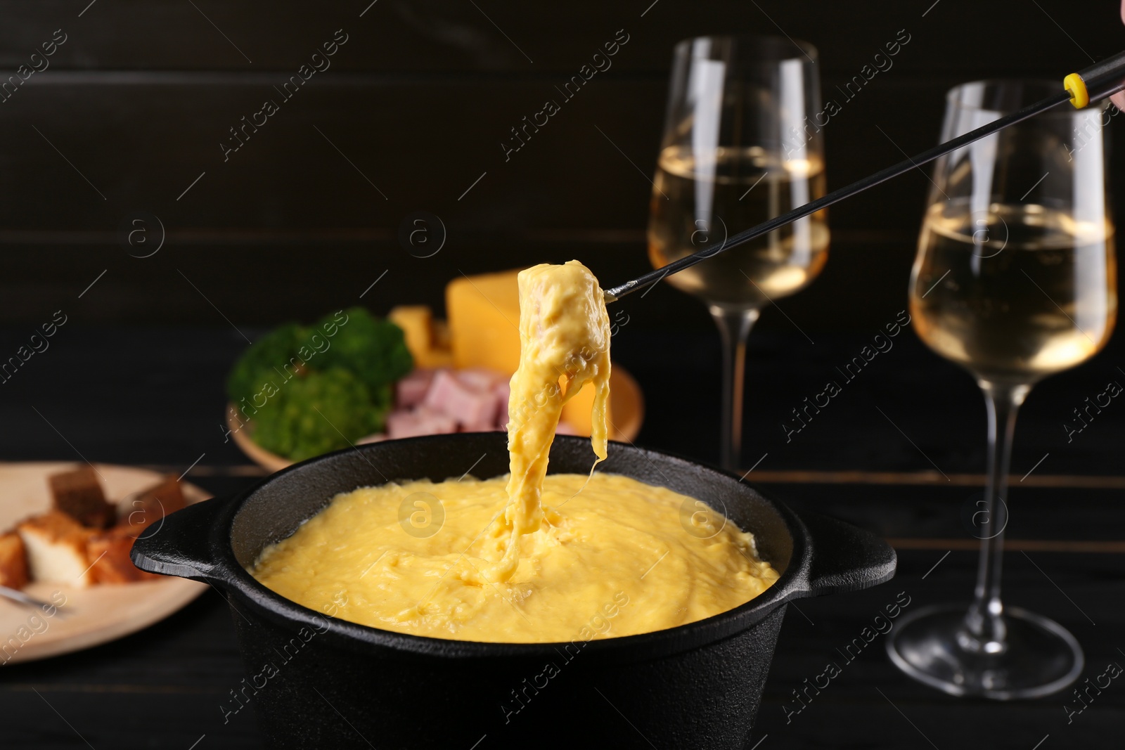 Photo of Dipping piece of ham into fondue pot with melted cheese on table, closeup