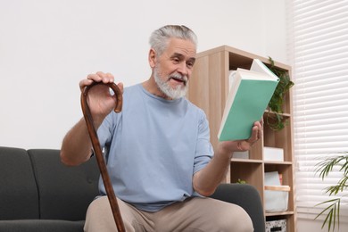 Photo of Senior man with walking cane reading book on sofa at home