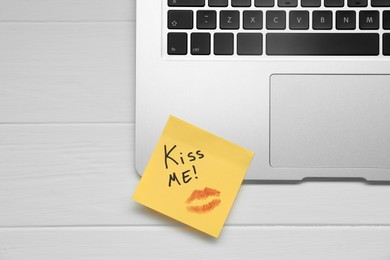 Photo of Sticky note with phrase Kiss Me and lipstick mark on laptop at white wooden table, top view