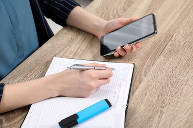 Woman taking notes while using smartphone at wooden table, closeup