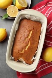 Photo of Tasty lemon cake in baking dish and citrus fruits on grey table, flat lay