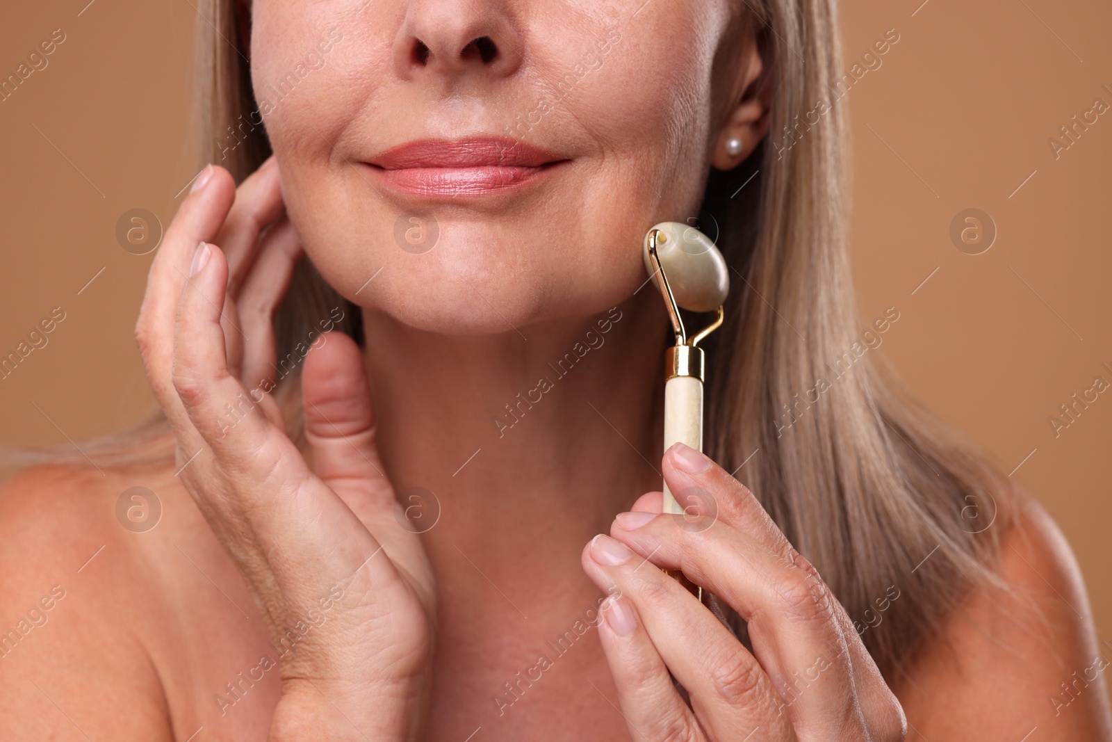 Photo of Woman massaging her face with jade roller on brown background, closeup