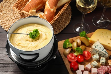 Photo of Fork with piece of broccoli, melted cheese in fondue pot, wine and snacks on wooden table, closeup