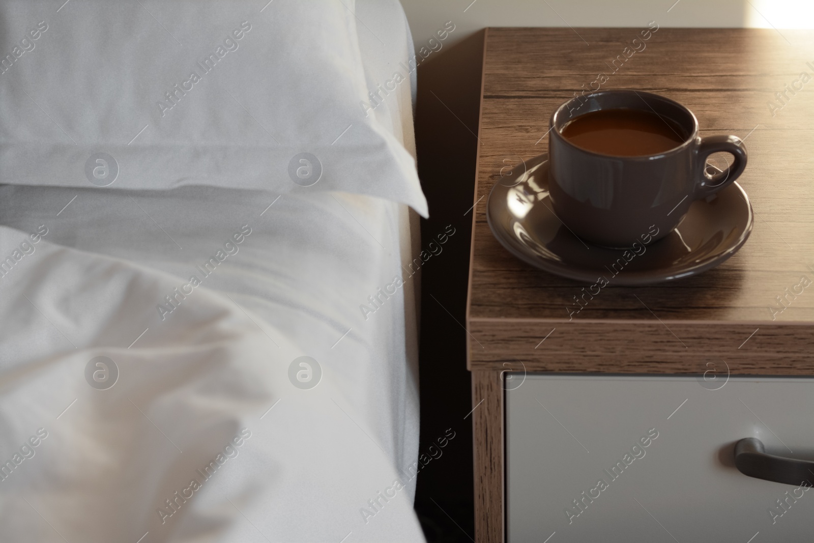 Photo of Cup of morning coffee on wooden night stand near bed indoors