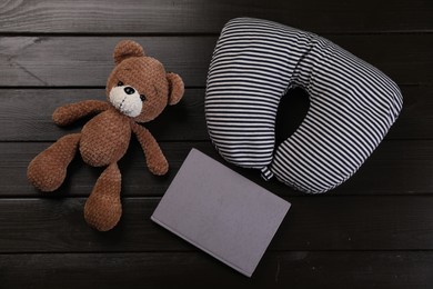 Photo of Striped travel pillow, notebook and toy bear on dark wooden background, flat lay