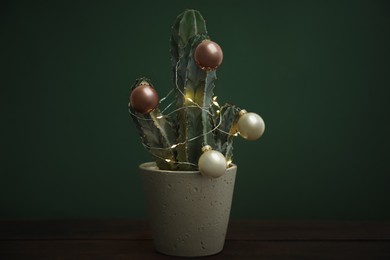 Beautiful cactus with Christmas balls and festive lights  on wooden table against green background