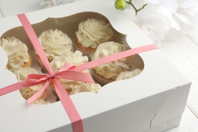 Photo of Tasty cupcakes with vanilla cream in box and orchid flowers on white wooden table, closeup