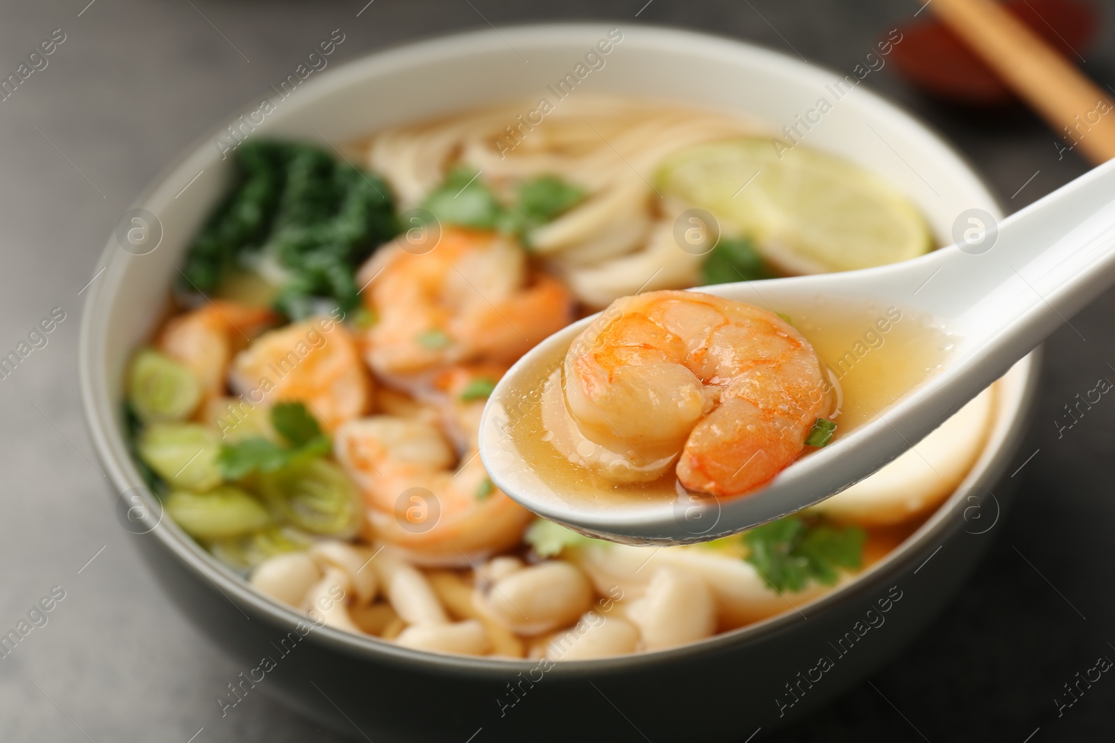 Photo of Eating delicious ramen with shrimps from bowl with spoon at grey table, closeup. Noodle soup