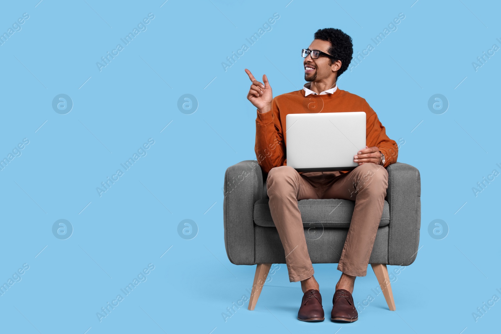 Photo of Happy man with laptop sitting in armchair and pointing at something on light blue background. Space for text