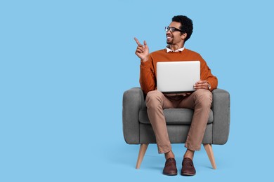 Happy man with laptop sitting in armchair and pointing at something on light blue background. Space for text