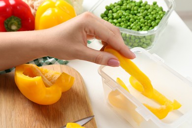 Woman putting cut bell pepper into plastic container at white table, closeup. Food storage