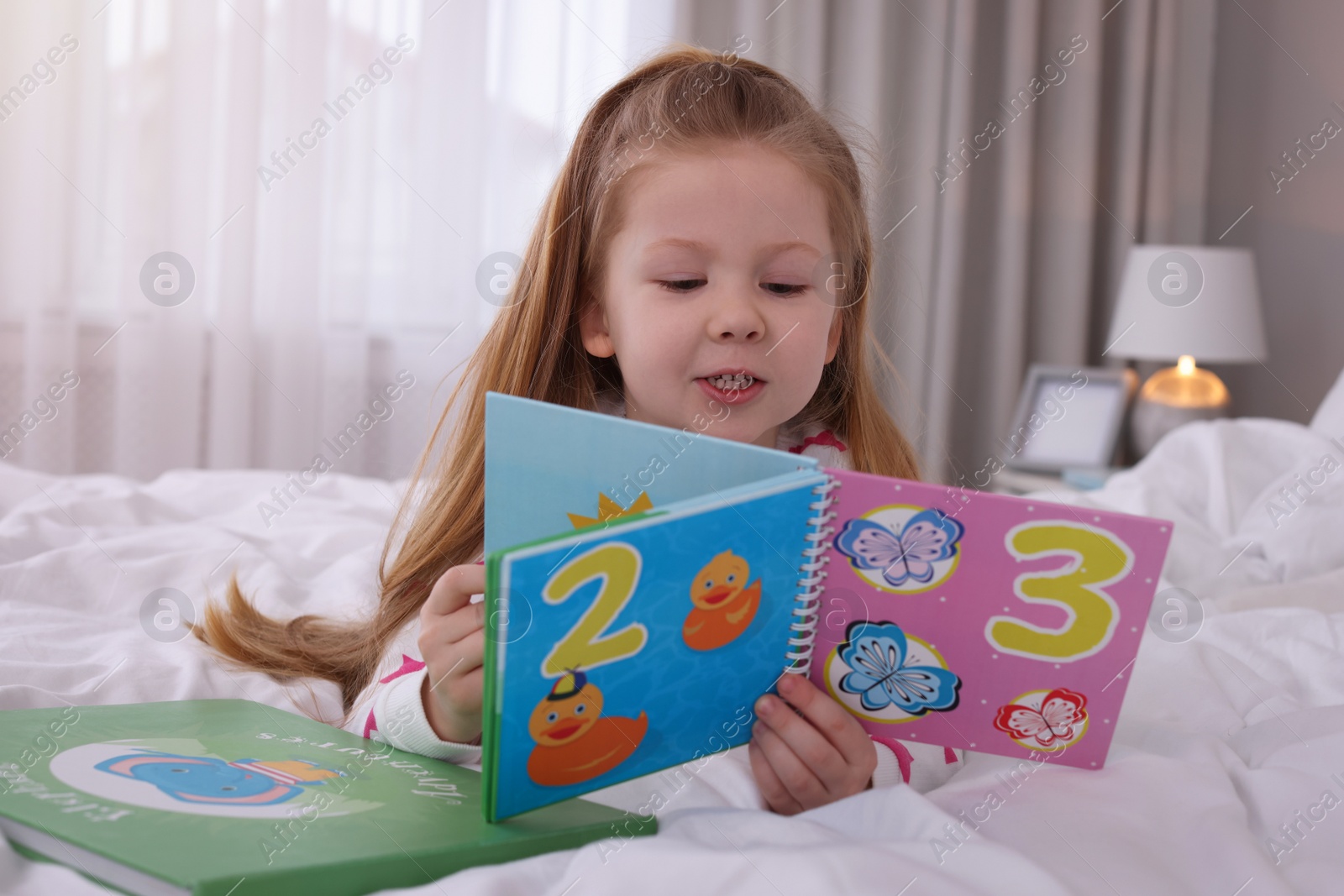 Photo of Cute little girl reading book on bed at home