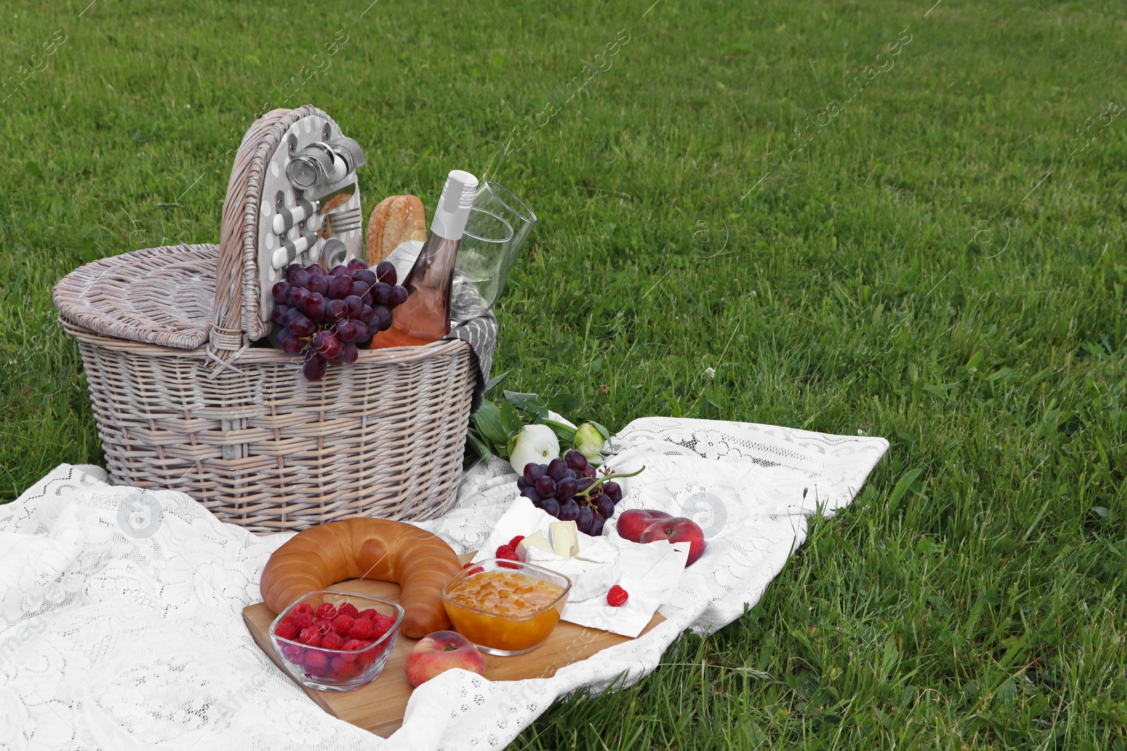 Photo of Picnic blanket with tasty food, flowers, basket and cider on green grass outdoors. Space for text