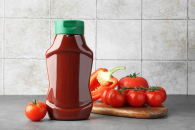 Photo of Bottle of tasty ketchup, tomatoes and pepper on grey table