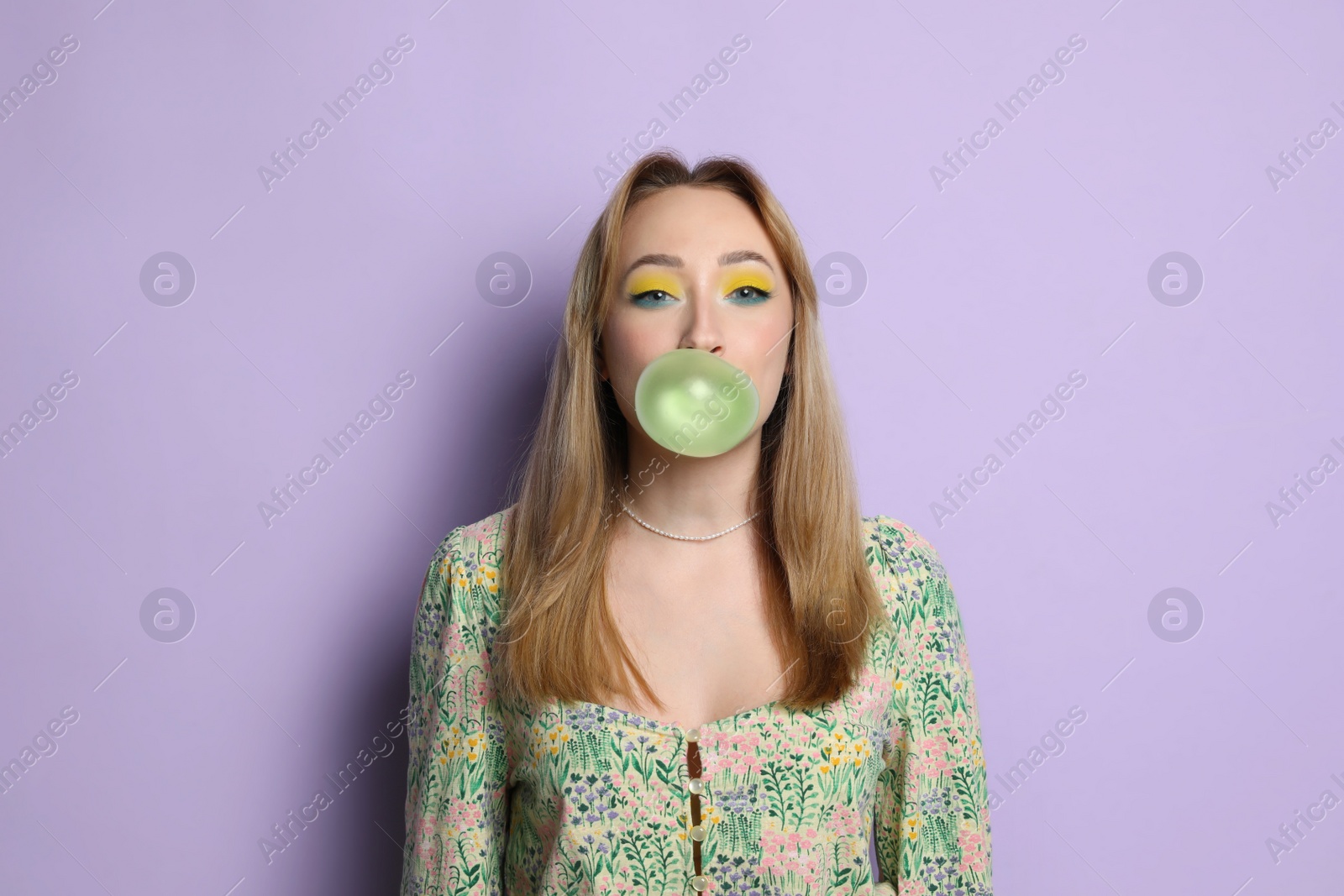 Photo of Fashionable young woman with bright makeup blowing bubblegum on lilac background