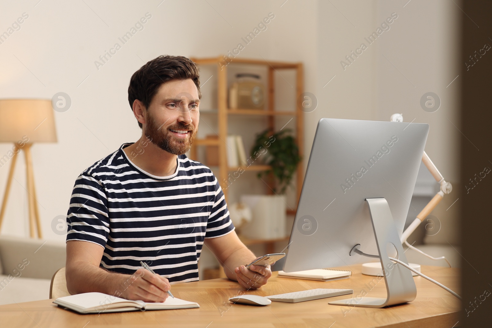 Photo of Home workplace. Happy man using smartphone and taking notes at wooden desk in room