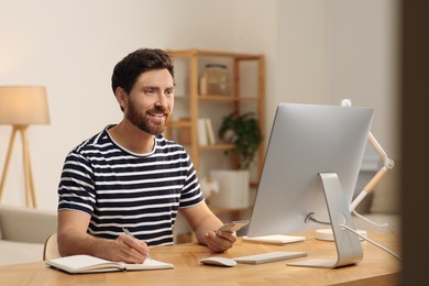 Home workplace. Happy man using smartphone and taking notes at wooden desk in room