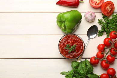 Delicious adjika sauce in bowl and ingredients on white wooden table, flat lay. Space for text