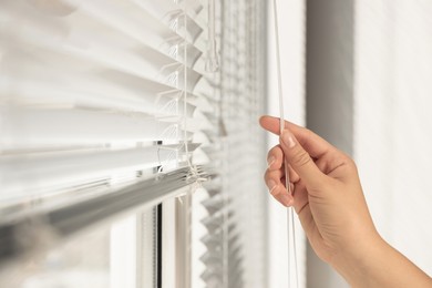 Woman opening horizontal blinds on window indoors, closeup