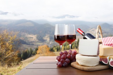 Photo of Different types of delicious cheeses, snacks and wine on wooden table against mountain landscape. Space for text