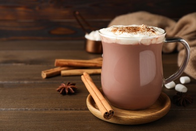 Photo of Glass cup of hot cocoa with whipped cream and aromatic cinnamon on wooden table. Space for text