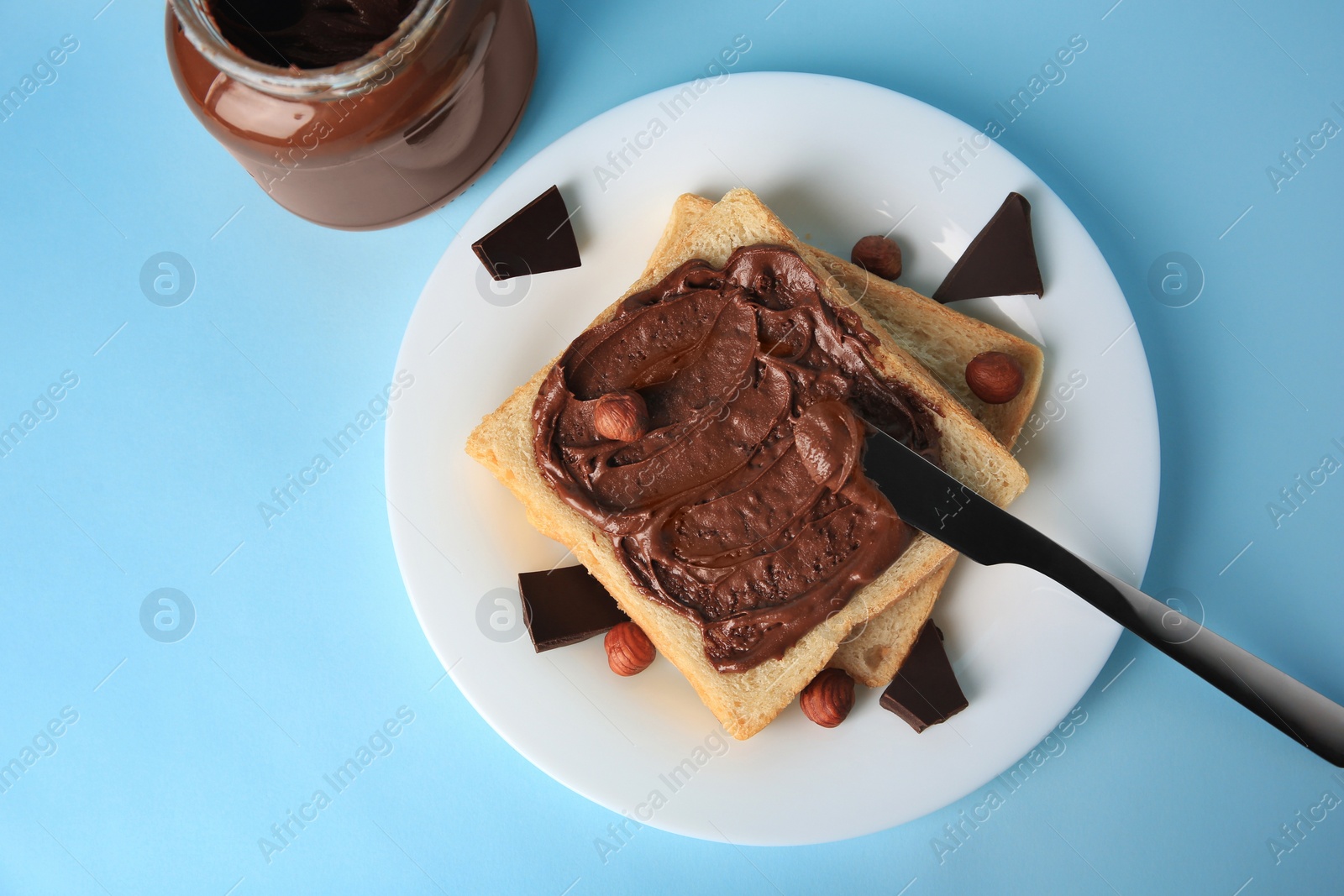 Photo of Spreading chocolate paste onto bread near jar of cream on light blue background, flat lay