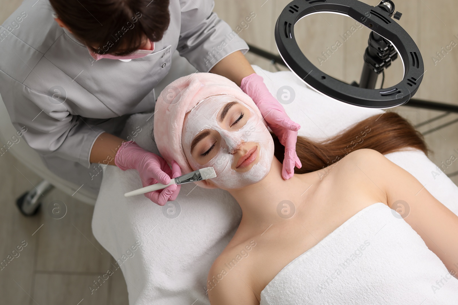 Photo of Cosmetologist applying mask on woman's face in clinic, top view