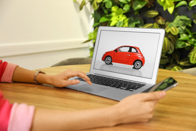 Photo of Woman using laptop and phone to buy car at wooden table indoors, closeup