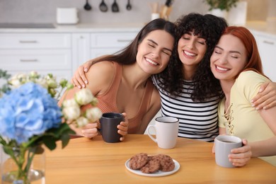Photo of Happy young friends with cups of drink spending time together at table in kitchen