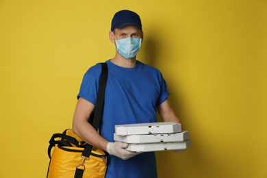 Photo of Courier in protective mask and gloves holding pizza boxes on yellow background. Food delivery service during coronavirus quarantine