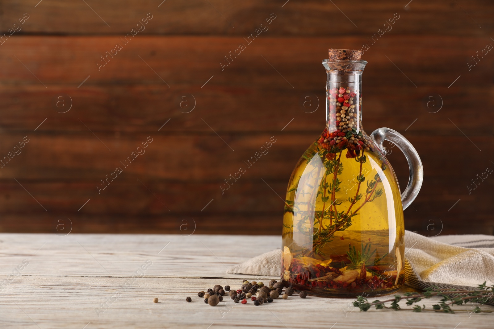 Photo of Glass jug of cooking oil with spices and herbs on white wooden table. Space for text