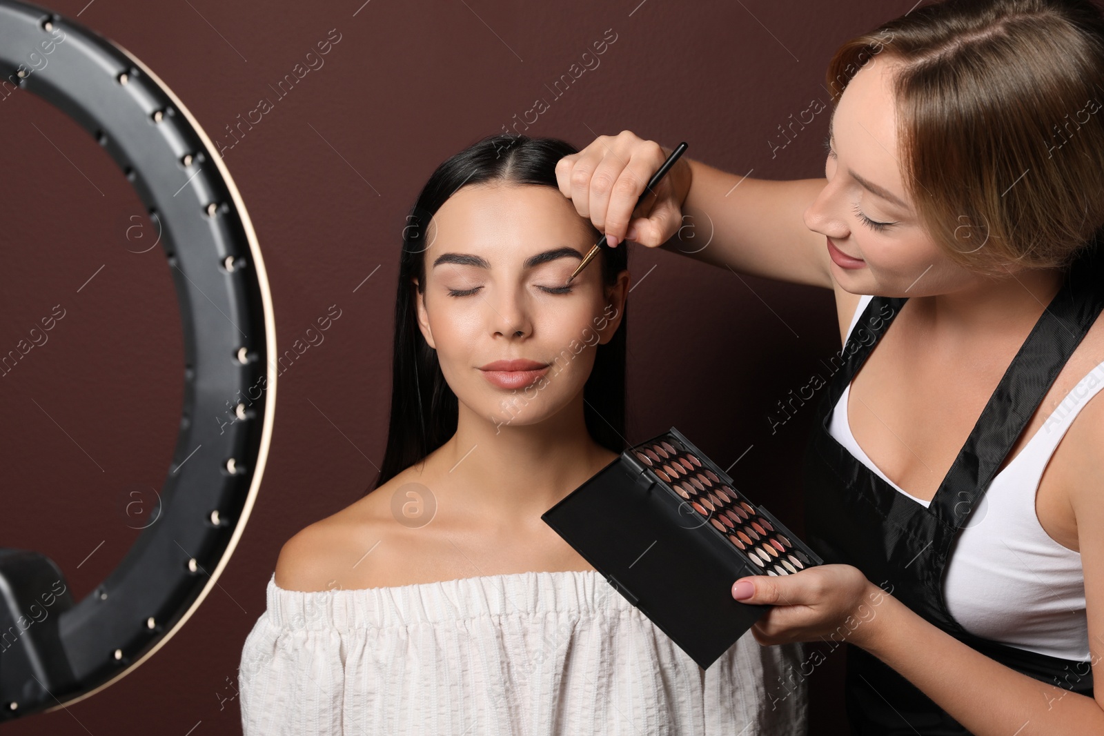 Photo of Professional makeup artist working with beautiful young woman against brown background. Using ring lamp