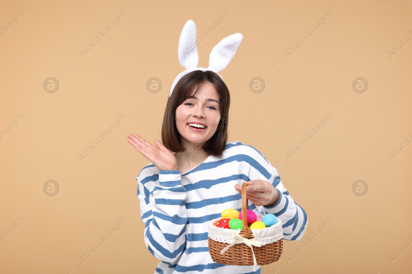 Photo of Easter celebration. Happy woman with bunny ears and wicker basket full of painted eggs on beige background