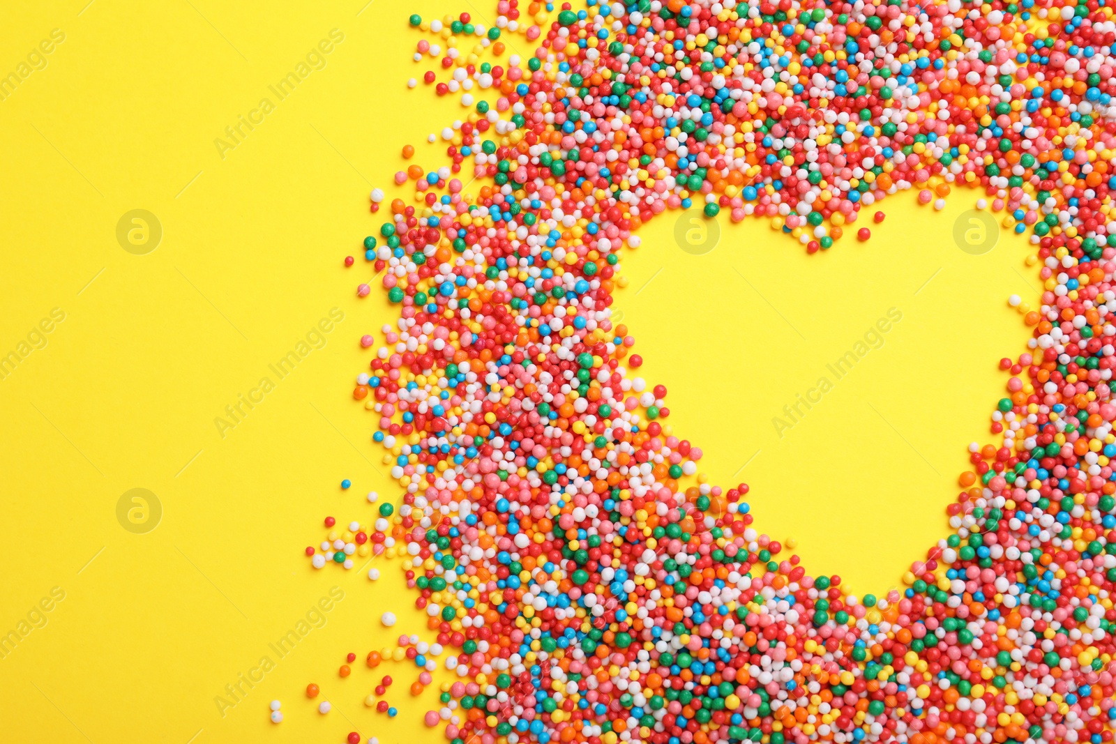 Photo of Heart shaped frame of sprinkles on yellow background, flat lay with space for text. Confectionery decor