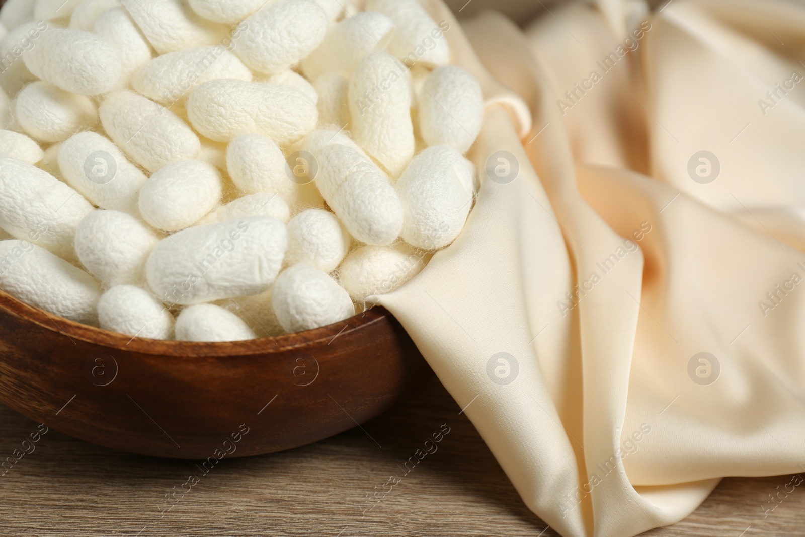 Photo of White cocoons with bowl and silk fabric on wooden table, closeup