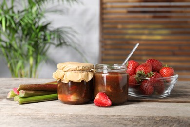 Jars of tasty rhubarb jam, fresh stems and strawberries on wooden table. Space for text
