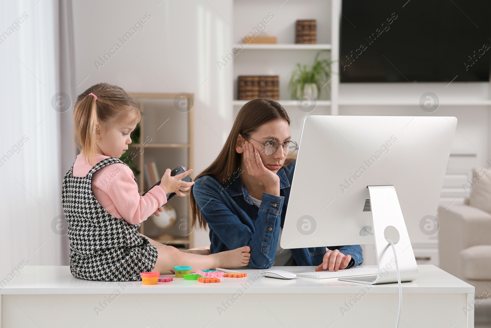 Photo of Woman working remotely at home. Tired mother using computer while her daughter playing with phone. Child sitting on desk